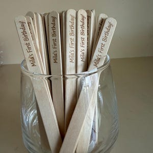 May include: A collection of light-colored wooden sticks held in a clear glass container. Each stick features the laser-engraved text "Mila's First Birthday!" in a dark brown script font, positioned vertically along the length of the wooden surface.