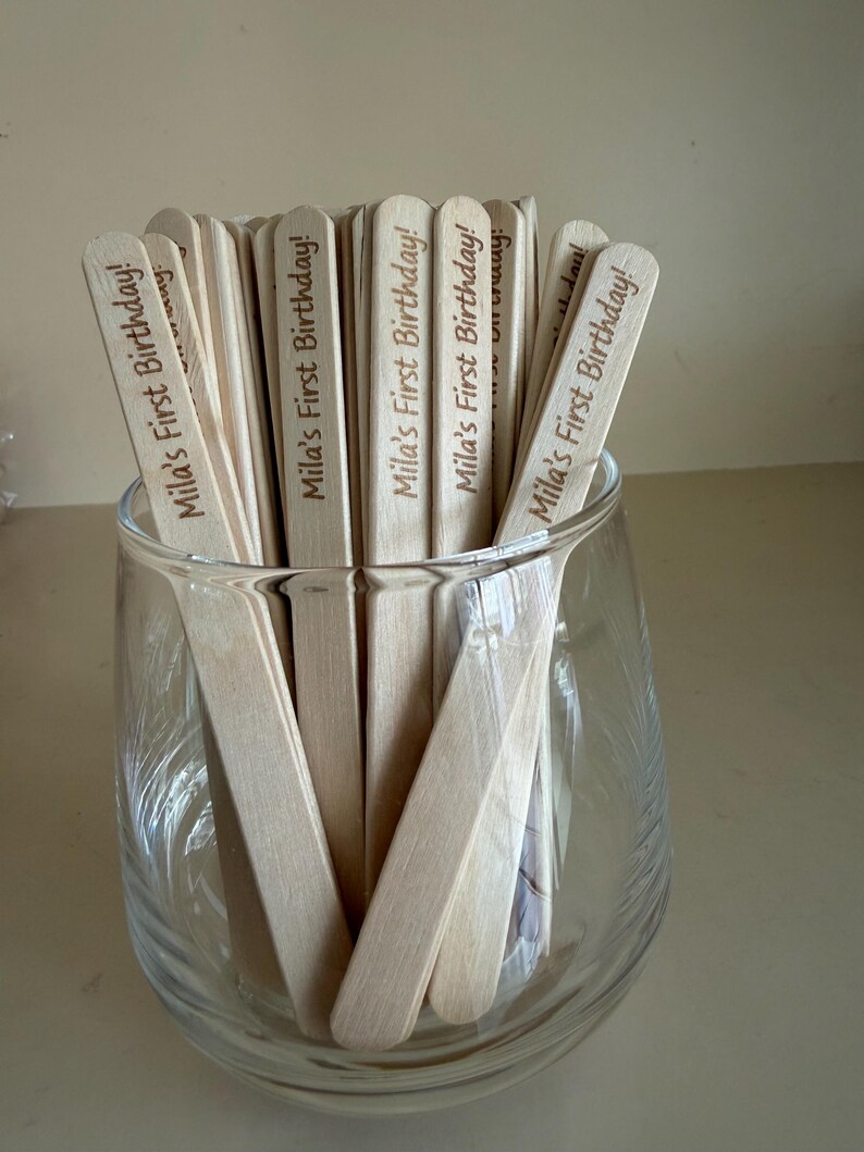 May include: A collection of light-colored wooden sticks held in a clear glass container. Each stick features the laser-engraved text "Mila's First Birthday!" in a dark brown script font, positioned vertically along the length of the wooden surface.
