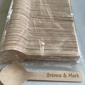 May include: A bundle of disposable wooden spoons in a clear plastic wrapper. One spoon is separate, with the engraved text "Brenna & Mark". The spoons are light brown and appear to be made of wood.