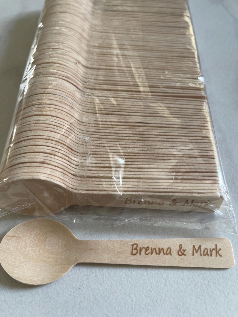 May include: A bundle of disposable wooden spoons in a clear plastic wrapper. One spoon is separate, with the engraved text "Brenna & Mark". The spoons are light brown and appear to be made of wood.