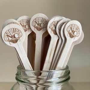 May include: A collection of light-colored wooden stirrers with round tops, each featuring a laser-engraved design of a seashell with the letters N&C centered inside. The stirrers are bundled together and placed upright inside a clear glass jar.