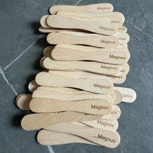 May include: A stack of light-colored wooden ice cream sticks, each featuring the word Magnuv branded in dark brown ink. The sticks are flat with rounded ends and are arranged in an overlapping pile, showcasing the natural wood grain texture.