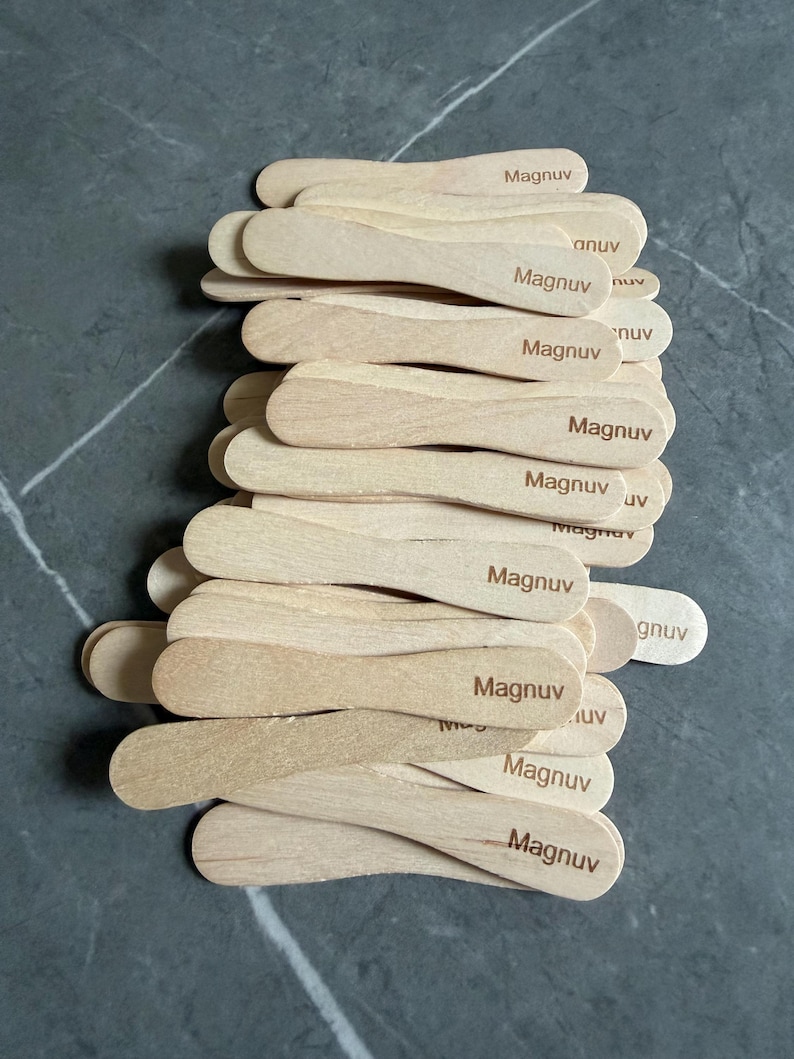 May include: A stack of light-colored wooden ice cream sticks, each featuring the word Magnuv branded in dark brown ink. The sticks are flat with rounded ends and are arranged in an overlapping pile, showcasing the natural wood grain texture.