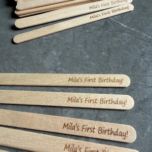May include: A collection of light-colored wooden craft sticks, each featuring the laser-engraved text "Mila's First Birthday!" in a simple, dark brown font. The sticks are arranged in a stack at the top and spread out individually below.