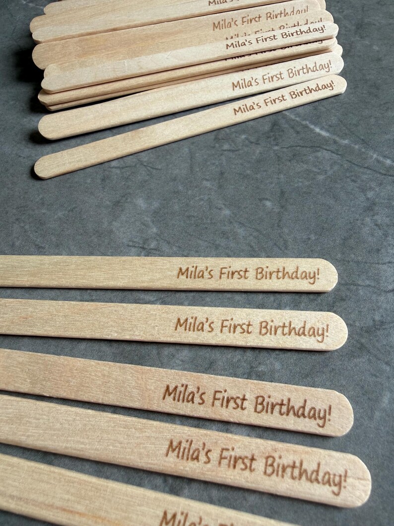 May include: A collection of light-colored wooden craft sticks, each featuring the laser-engraved text "Mila's First Birthday!" in a simple, dark brown font. The sticks are arranged in a stack at the top and spread out individually below.