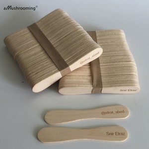 May include: Two stacks of light-colored wooden craft sticks bound by brown paper bands, with two loose sticks placed in front. The text aMushrooming, @efrat_abed, and Snir Elraz are printed on the wood surfaces in a dark brown font.