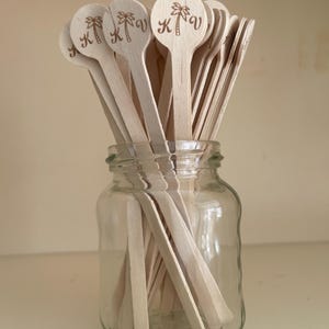 May include: A clear glass jar filled with wooden spoons. The spoons have a round head with a palm tree and the letters "K" and "V" engraved on them. The spoons are light brown and the jar is sitting on a white surface.