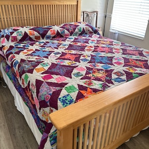 Op de afbeelding: Een houten bedframe met een wit en paars patchwork dekbed. Het dekbed heeft een stervormig patroon en is gemaakt van verschillende stoffen.