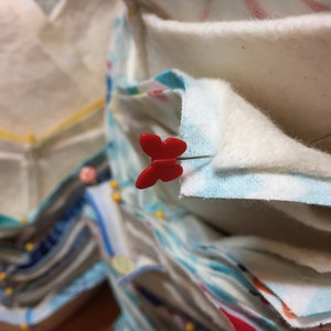 May include: A stack of fabric squares pinned together with red butterfly-shaped pins. The fabric is a variety of colors and patterns, including white, blue, and red.