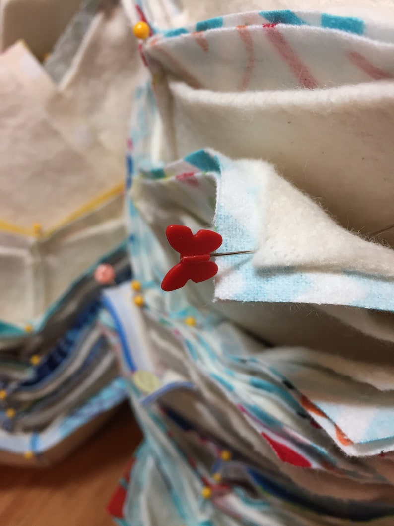 May include: A stack of fabric squares pinned together with red butterfly-shaped pins. The fabric is a variety of colors and patterns, including white, blue, and red.
