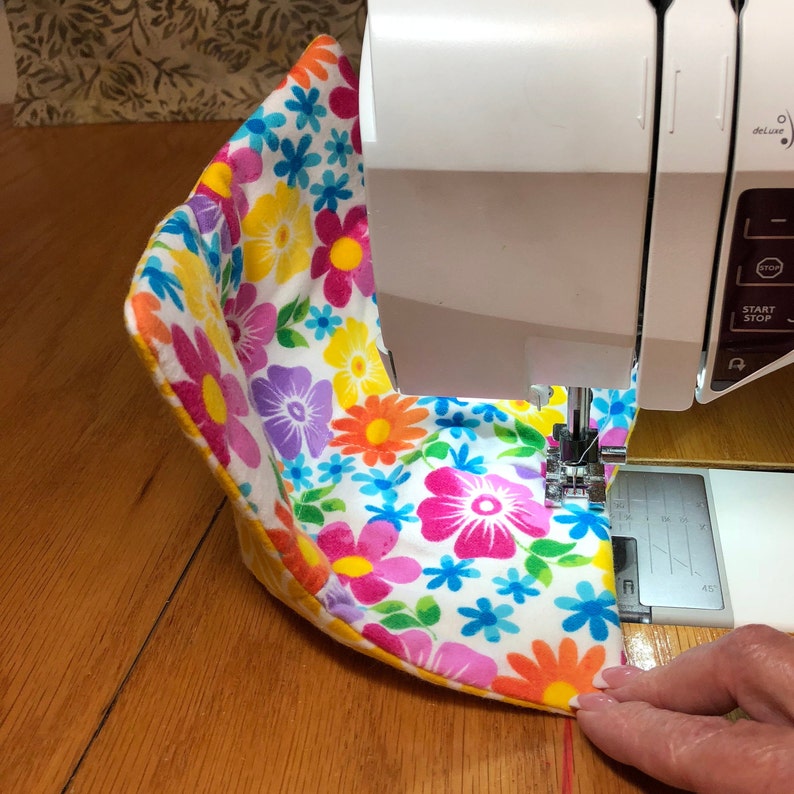 May include: A sewing machine with a floral fabric being sewn. The fabric is white with pink, yellow, blue, and orange flowers. The sewing machine is white with a black control panel.