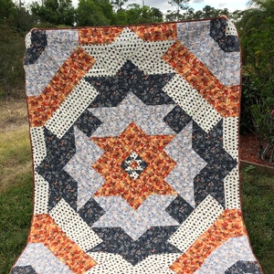 Op de afbeelding: Een groot quilt met een stervormig patroon in tinten oranje, bruin, wit en blauw. Het quilt is gemaakt van verschillende stoffen met verschillende patronen, waaronder stippen, bloemenprints en geometrische ontwerpen.