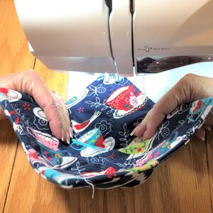 May include: A person is sewing a blue fabric with a pattern of teacups and flowers on a sewing machine. The fabric is folded in half and the person is holding the fabric in place with their hands.