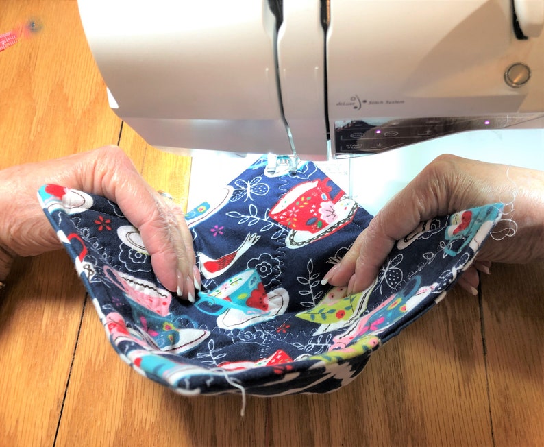 May include: A person is sewing a blue fabric with a pattern of teacups and flowers on a sewing machine. The fabric is folded in half and the person is holding the fabric in place with their hands.