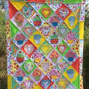 May include: A colorful quilt with a patchwork design featuring cupcakes and hearts. The quilt is made with a variety of fabrics in shades of pink, yellow, green, blue, and white.