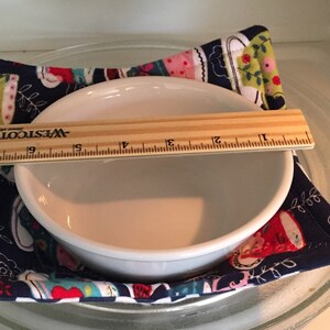 May include: A white ceramic bowl sits on a blue and white fabric hot pad with a teacup pattern. A wooden ruler with the text "WESTCOTT" is placed across the top of the bowl.