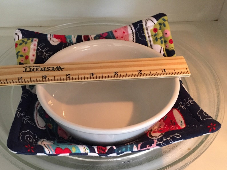 May include: A white ceramic bowl sits on a blue and white fabric hot pad with a teacup pattern. A wooden ruler with the text "WESTCOTT" is placed across the top of the bowl.