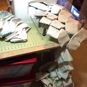 May include: A white sewing machine with a clear plastic sewing table. The table has a grid pattern and is covered with beige fabric squares. A stack of beige fabric squares with blue and white trim is on the left side of the table. A stack of beige fabric squares with blue and white trim is on the floor.