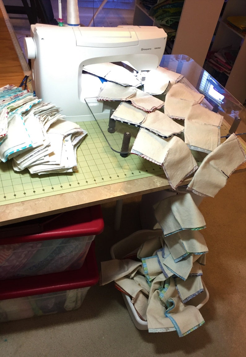 May include: A white sewing machine with a clear plastic sewing table. The table has a grid pattern and is covered with beige fabric squares. A stack of beige fabric squares with blue and white trim is on the left side of the table. A stack of beige fabric squares with blue and white trim is on the floor.