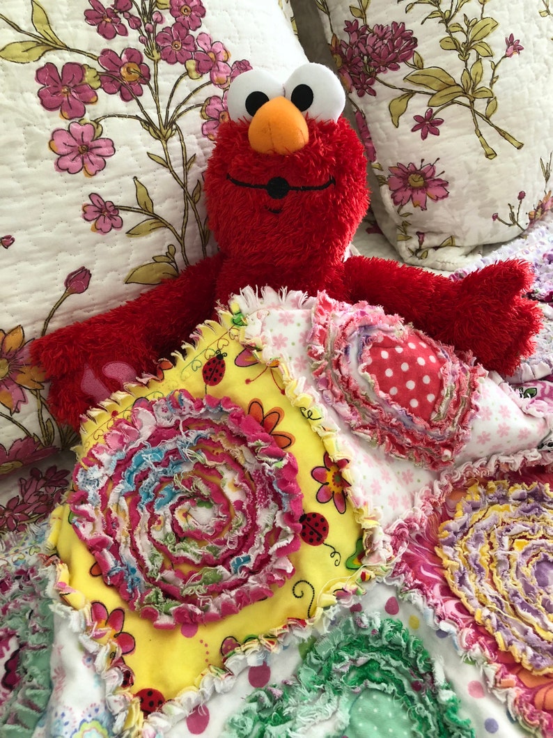 Puede incluir: Un peluche rojo de Elmo est&aacute; acostado sobre una colcha de retazos colorida hecha a mano. La colcha est&aacute; hecha de varias telas con diferentes patrones y texturas, incluyendo flores, lunares y remolinos. La colcha es una adici&oacute;n brillante y alegre a la habitaci&oacute;n de cualquier ni&ntilde;o.