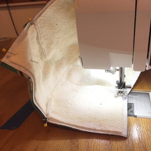 May include: A close-up of a sewing machine stitching a white fabric with a green and white striped fabric underneath. The fabric is pinned in place with yellow pins.