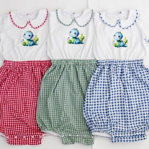 Puede incluir: Tres peleles para bebés con tops blancos y bajos de cuadros. Cada pelele presenta un cuello Peter Pan con ribete a juego. Los tops tienen una aplicación de dinosaurio y cactus. Los bajos son de cuadros rojos, verdes y azules.