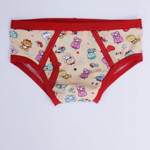 May include: Briefs with a cream-colored base featuring a repeating pattern of colorful cartoon bears, small hearts, and stars. The garment has a solid red elastic waistband and red trim along the leg openings and the front center panel.