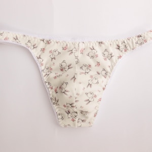 Könnte beinhalten: Cremefarbener String-Tanga mit einem sich wiederholenden Muster aus Cartoon-Hasen und rosa Blumen. Die Kanten sind mit weißer Spitze verziert. Der Stoff scheint ein weiches, leichtes Material zu sein.