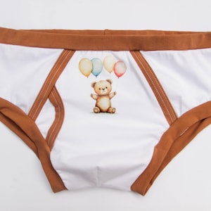 Puede incluir: Ropa interior blanca con ribetes y detalles marrones. La parte delantera presenta una ilustración de acuarela de un oso de peluche con globos. La ropa interior tiene una cintura marrón y detalles laterales marrones.