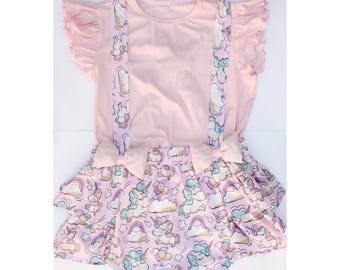 May include: A light pink romper set with a matching headband. The romper has a ruffled top, suspenders, and a skirt decorated with a unicorn and cloud pattern. The headband features a bow with a star design.