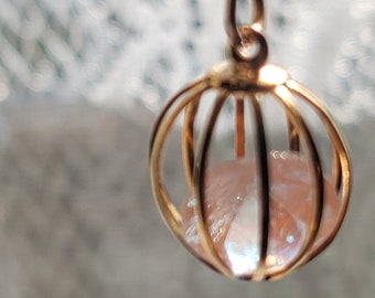 Pode incluir: Um colar com pingente dourado com um design de gaiola esférica. Dentro da gaiola há uma pedra rosa claro iridescente. A corrente do colar também é dourada e o fundo está desfocado.