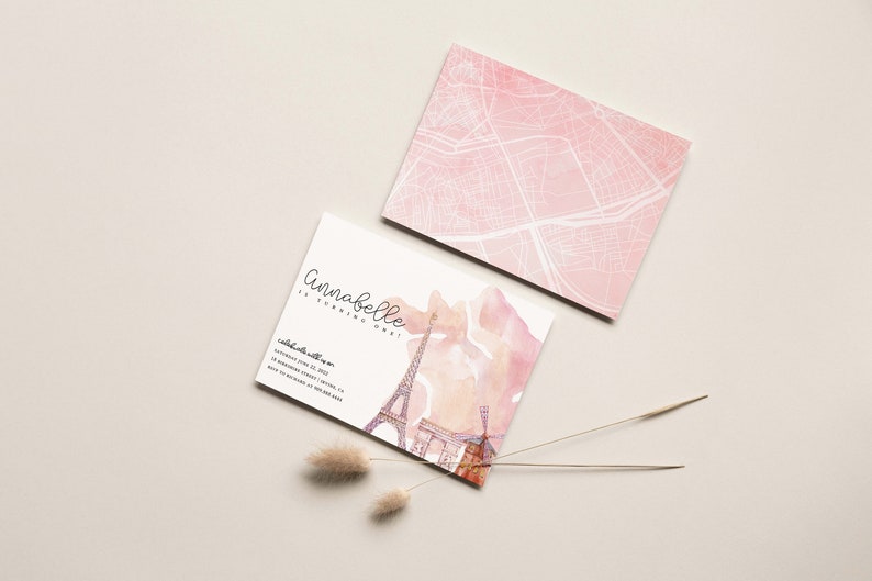 以下が含まれることがあります： ピンクと白の招待状。水彩画で描かれたエッフェル塔のイラストと「アナベルは1歳になります！」というテキストが書かれています。