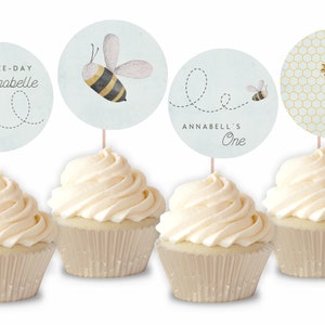 May include: Four cupcakes with white frosting topped with round, blue and white bee-themed cupcake toppers. The toppers feature a bee, honeycomb, and the text "Happy Bee-Day Annabelle" and "Annabell's One".