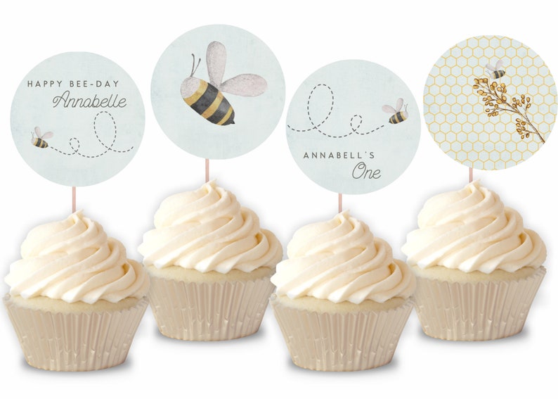 May include: Four cupcakes with white frosting topped with round, blue and white bee-themed cupcake toppers. The toppers feature a bee, honeycomb, and the text "Happy Bee-Day Annabelle" and "Annabell's One".