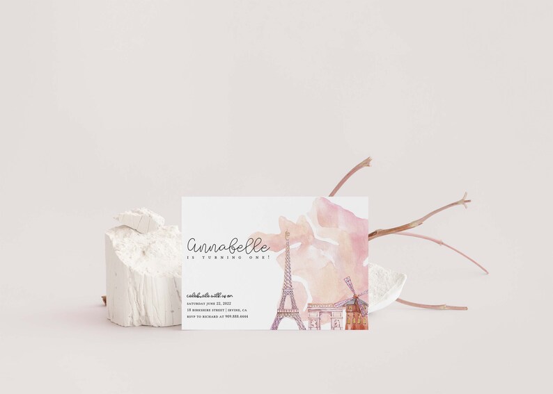 May include: A pink and white watercolor invitation with a Parisian theme. The invitation says "Annabelle is turning one!" and includes a watercolor illustration of the Eiffel Tower, Arc de Triomphe, and a windmill.