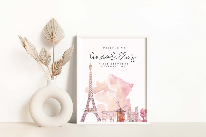 K&ouml;nnte beinhalten: Ein wei&szlig;er gerahmter Druckbarer mit einer Aquarell-Illustration des Eiffelturms, einer Windm&uuml;hle und einer Kirche. Der Text auf dem Schild lautet "Willkommen zur ersten Geburtstagsfeier von Annabelle."