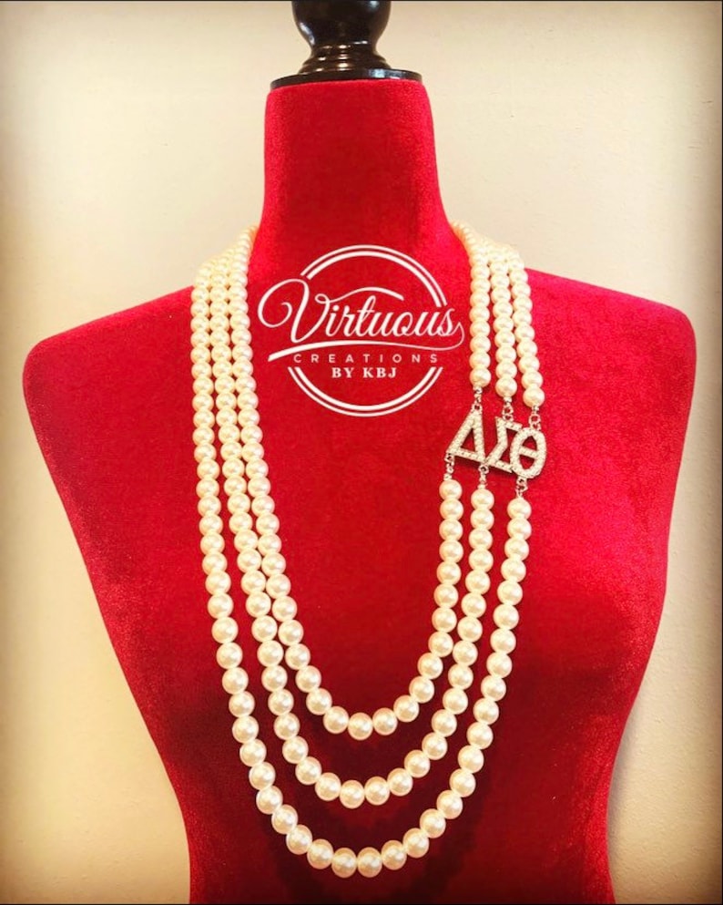 Puede incluir: Un collar de perlas de tres hileras con un dije dorado que dice "&Delta;&Sigma;&Theta;" en pedrer&iacute;a. El collar est&aacute; en un maniqu&iacute; de terciopelo rojo.