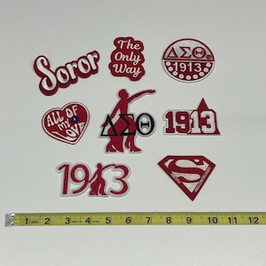 May include: A collection of embroidered patches in red and white, featuring various designs and text. The patches include phrases like "Soror" and "The Only Way," along with Greek letters and the number "1913." A measuring tape is included.