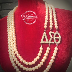 Puede incluir: Un collar de perlas de tres hilos con un dije de plata con las letras griegas Delta Sigma Theta. El collar se muestra sobre un fondo de terciopelo rojo.