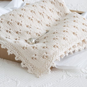 May include: A cream-coloured crocheted baby blanket with a delicate lace border. The blanket is folded and placed in a cardboard box, with a silver pin on top.