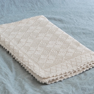 May include: A cream-colored crocheted throw blanket with a diamond pattern and a crocheted border. The blanket is lying on a light blue surface.