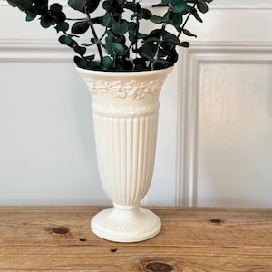 May include: A cream-colored ceramic vase with a flared rim, vertical fluted detailing on the body, and a raised floral relief pattern around the neck. The vase is filled with several tall, dark green eucalyptus branches with small round leaves.