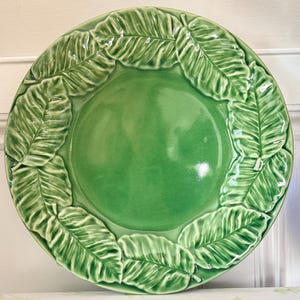 May include: A round ceramic plate with a glossy green finish. The center of the plate is smooth and concave, while the wide rim features a raised, embossed pattern of overlapping leaves that circle the entire perimeter of the dish.