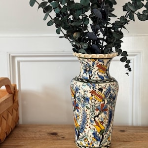 May include: A ceramic vase with a scalloped rim, featuring a cream-colored base decorated with intricate hand-painted floral patterns in shades of blue, yellow, and orange, with dark green foliage accents and black outlines throughout the design.