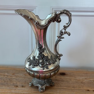 May include: A silver-toned metal pitcher featuring an ornate, curved handle and a scalloped rim. The body is decorated with embossed floral motifs and a fluted base, standing on three small feet with a polished, metallic finish throughout.