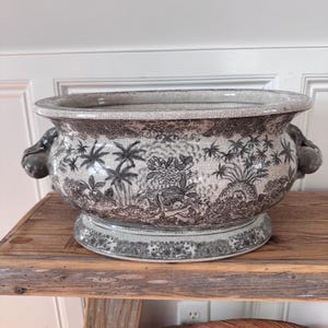 Può includere: Un vaso da fiori in ceramica con finitura smaltata a craquelé, caratterizzato da un motivo a trasferimento in bianco e nero di palme tropicali, fogliame e piccole figure. Il vaso ovale ha un bordo largo, una base a piedistallo e due manici.