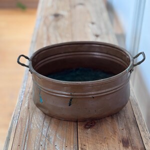 May include: A round, metallic copper basin featuring two small, curved handles on opposite sides. The exterior surface displays a warm, weathered brown tone with subtle hints of blue oxidation and minor indentations along the lower rim.