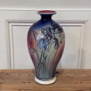 May include: A ceramic vase with a flared rim and a tapered base, featuring a hand-painted iris flower design in shades of blue, green, and purple. The background transitions from a deep blue at the top to a soft mauve and pink hue below.