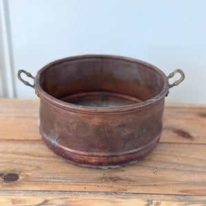 May include: A round copper basin with a weathered, dark metallic finish and visible green oxidation. The vessel features two small, curved brass handles attached to opposite sides of the rim, with a smooth, cylindrical body and a flat base.