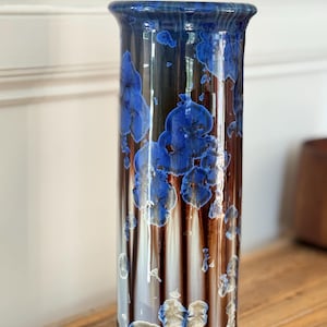 Könnte beinhalten: Eine hohe, zylindrische Keramikvase mit glänzender Oberfläche und einer tiefbraunen Grundfarbe. Die Oberfläche ist mit kristallinen Glasurmustern in leuchtenden Blau- und Weißtönen verziert, die ausgeprägte blumenartige Formen bilden.